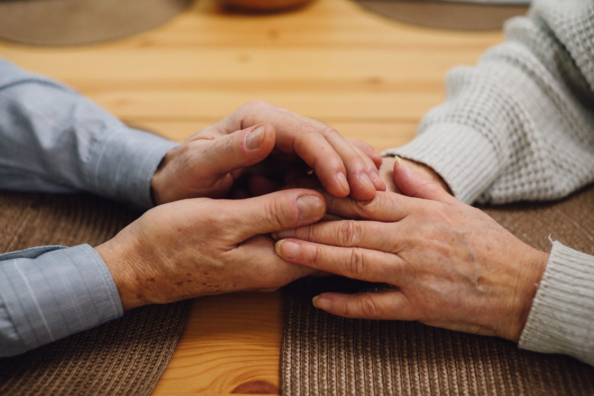 An older couple's hands clasped together on a wooden table