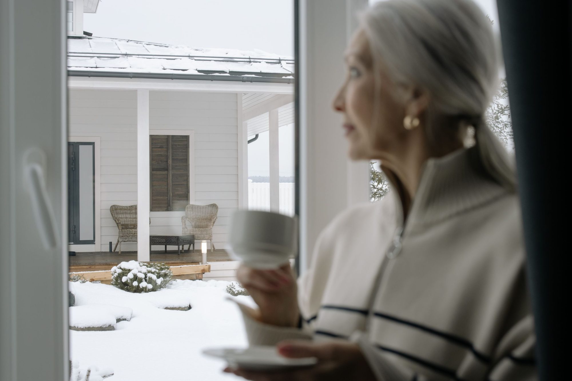 An older woman with a mug beside a bright window on an ordinary morning